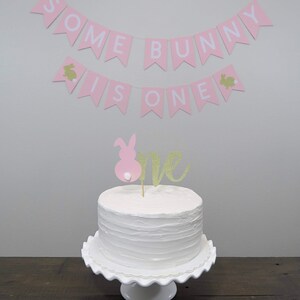 May include: A white cake with pink and gold glitter frosting and a pink bunny topper. The cake is on a white cake stand. A pink banner with the text "Some Bunny Is One" hangs above the cake.