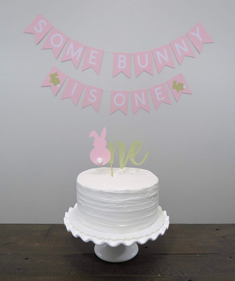May include: A white cake with pink and gold glitter frosting and a pink bunny topper. The cake is on a white cake stand. A pink banner with the text "Some Bunny Is One" hangs above the cake.