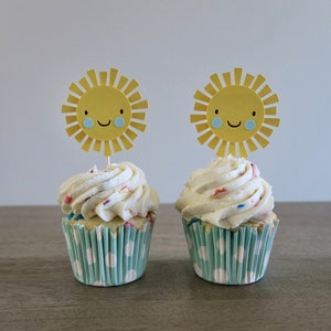 May include: Two cupcakes with white frosting and colorful sprinkles topped with yellow paper sun toppers with blue smiling faces.