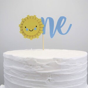 May include: A white cake with a blue and yellow cake topper that says "one" with a smiling sun with blue eyes.