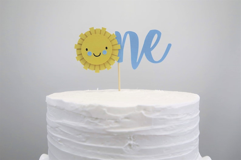 May include: A white cake with a blue and yellow cake topper that says "one" with a smiling sun with blue eyes.