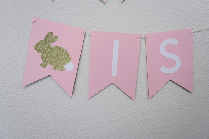 May include: Pink and white banner with the word "It's" and a gold glitter bunny with a white tail.