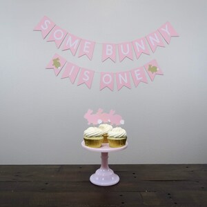 May include: Pink and white banner with the text "Some Bunny Is One" hanging on a white wall. Three cupcakes with white frosting and bunny toppers are on a pink cake stand.