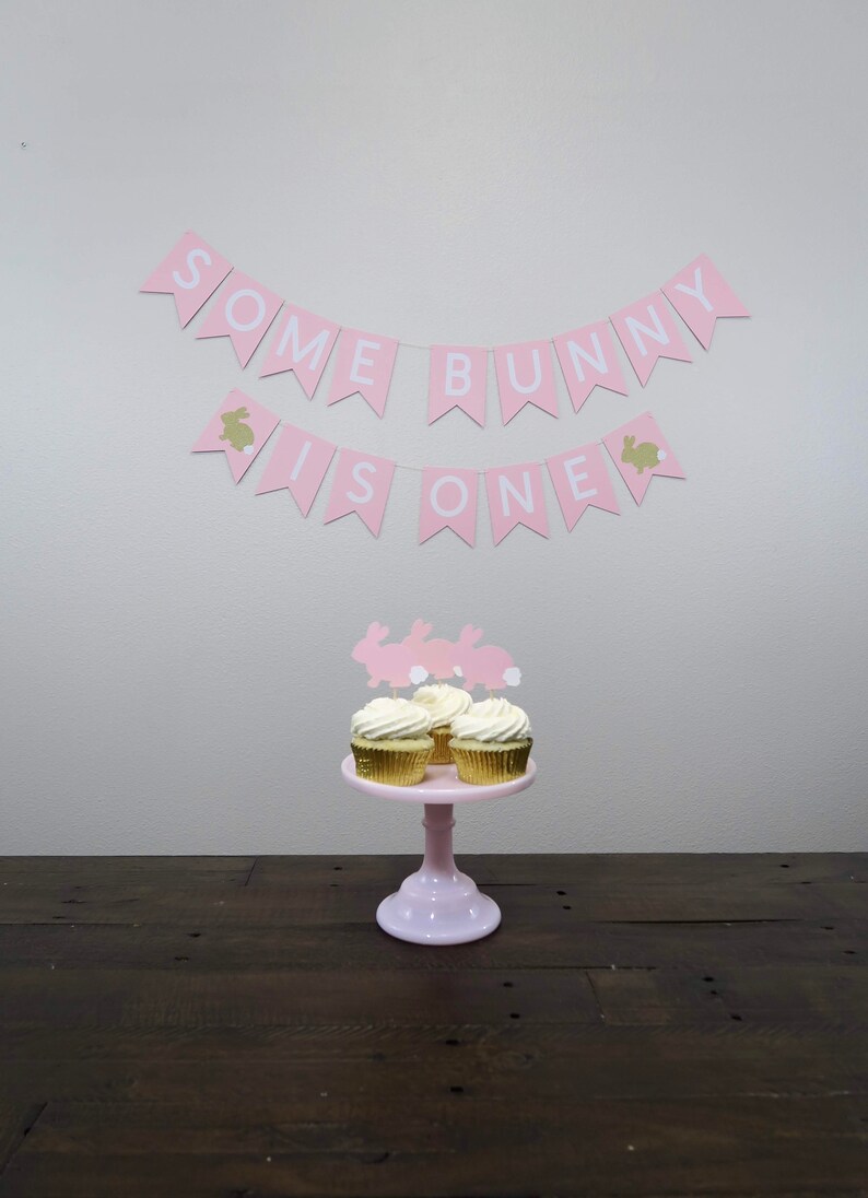 May include: Pink and white banner with the text "Some Bunny Is One" hanging on a white wall. Three cupcakes with white frosting and bunny toppers are on a pink cake stand.