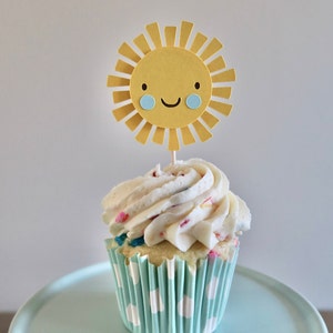 May include: A white cupcake with colorful sprinkles topped with white frosting and a yellow paper sun with a smiling face.