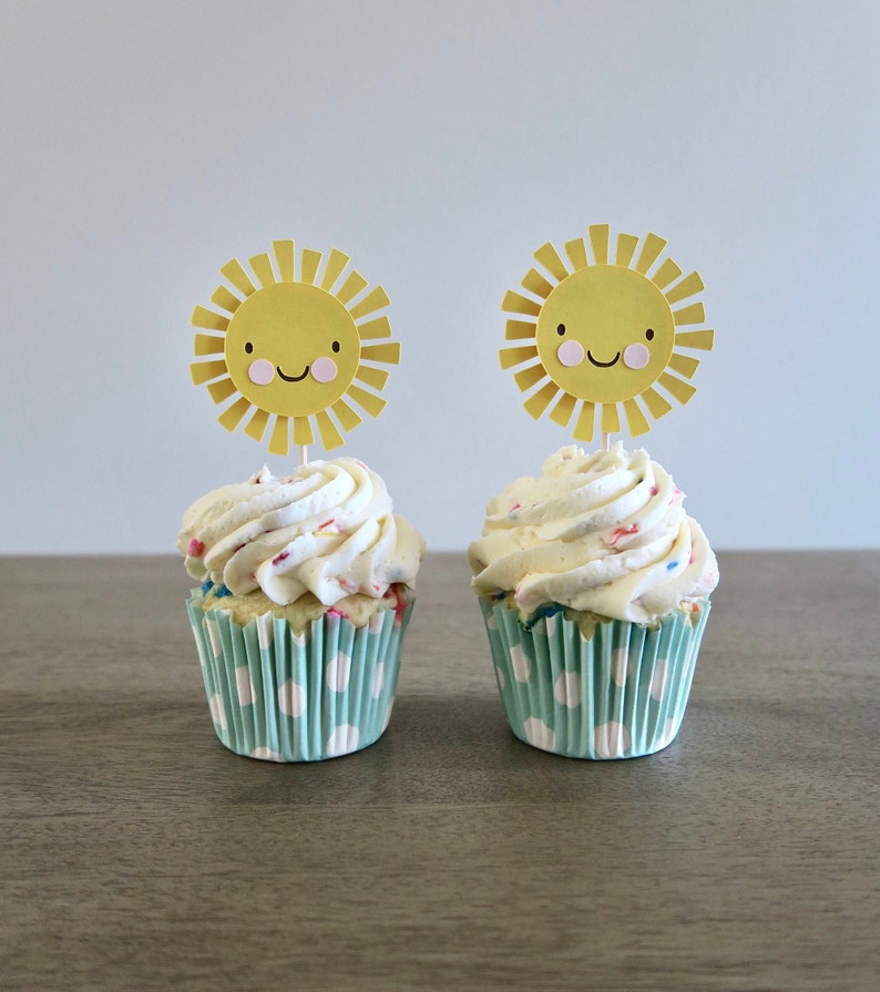 May include: Two white cupcakes with yellow and pink sprinkles topped with yellow paper sun toppers with smiling faces. The cupcakes are in blue and white polka dot liners.