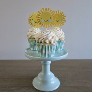 May include: Three cupcakes with white frosting and colorful sprinkles topped with yellow paper sun cutouts on a light blue cake stand.