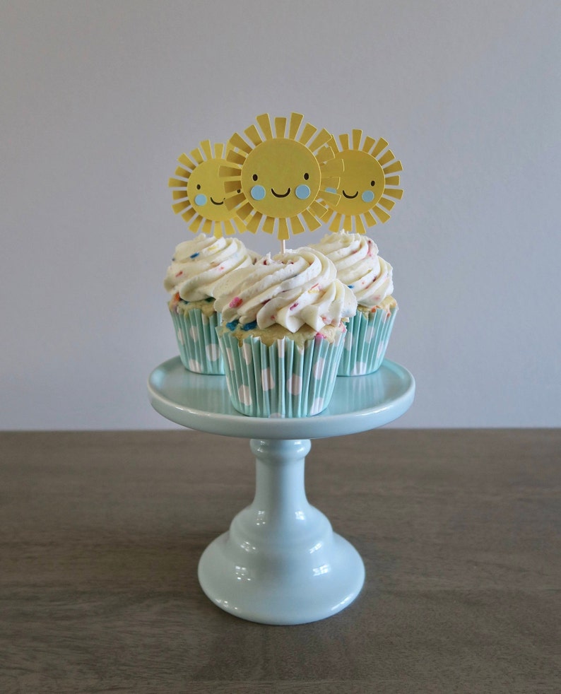May include: Three cupcakes with white frosting and colorful sprinkles topped with yellow paper sun cutouts on a light blue cake stand.