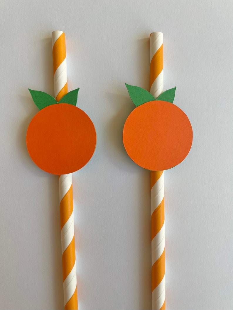 Peut inclure: Deux pailles en papier ray&eacute;es orange et blanc avec des cercles en papier orange et des feuilles en papier vert attach&eacute;s au sommet. Les cercles et les feuilles sont en forme d'orange.