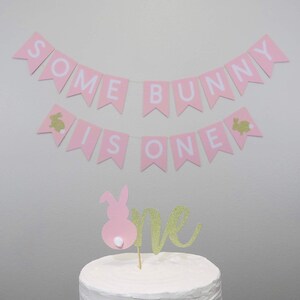 May include: Pink and white banner with the text "Some Bunny Is One" and gold bunny silhouettes. A pink bunny cake topper with a gold glitter "one" on a white frosted cake.