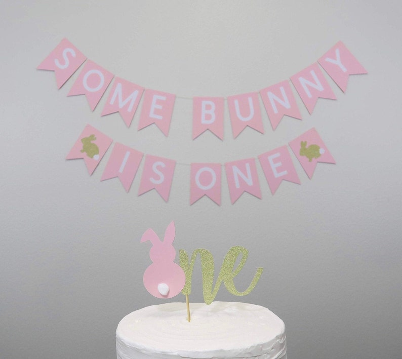 May include: Pink and white banner with the text "Some Bunny Is One" and gold bunny silhouettes. A pink bunny cake topper with a gold glitter "one" on a white frosted cake.
