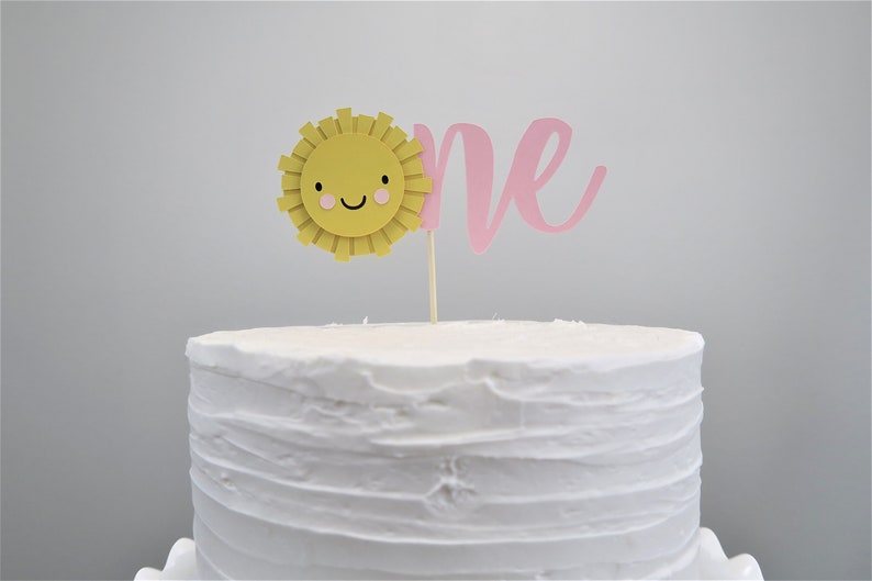 May include: A white cake with a pink "one" and a yellow sun with a smiling face on top. The sun has pink cheeks.
