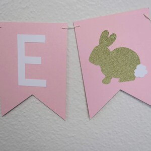 May include: Pink and white banner with the letter "E" and a gold glitter bunny with a white fluffy tail.
