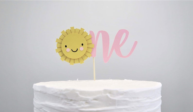 May include: A white cake with a yellow sun and pink "one" topper. The sun has a smiling face with pink cheeks.
