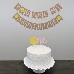May include: A white cake with pink and yellow frosting and a sun topper. A banner with the text "You are my sunshine" hangs above the cake.