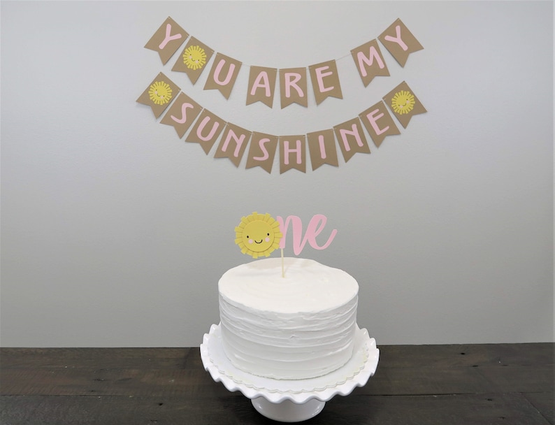 May include: A white cake with pink and yellow frosting and a sun topper. A banner with the text "You are my sunshine" hangs above the cake.