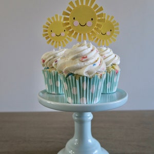 May include: Three cupcakes with white frosting and colorful sprinkles topped with yellow paper sun cutouts on a light blue cake stand.