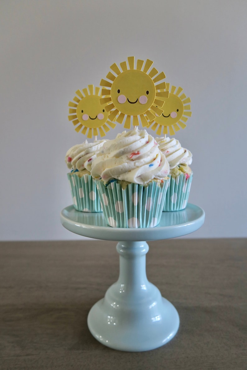 May include: Three cupcakes with white frosting and colorful sprinkles topped with yellow paper sun cutouts on a light blue cake stand.