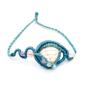 May include: A macrame bracelet crafted from teal cord featuring a central white seashell encased in woven knots. The design includes small, round, multi-colored beads in shades of purple, pink, green, and translucent white integrated into the cord.