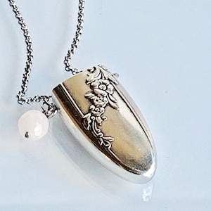 May include: Silver-toned pendant necklace featuring a floral design on a spoon-shaped pendant. The pendant hangs from a delicate chain and includes a small, round, off-white bead. The necklace is displayed against a light background.