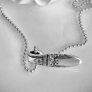 May include: A silver spoon pendant necklace. The pendant is a repurposed spoon with intricate floral and scrollwork details. The necklace has a delicate ball chain. The background is a white, textured surface.
