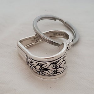 May include: Silver-toned spoon handle repurposed as a keychain. The spoon handle features a floral design around the edge and is attached to a silver key ring. The background is a white, textured surface.