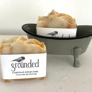 May include: Two bars of artisan soap, one in a small gray bathtub-shaped holder. The soap is wrapped in white paper with the word "grounded" and a bird illustration. The soap is a light brown color and the net weight is 4 oz (113 g).