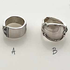 May include: Two silver rings made from repurposed silverware. The ring on the left has a simple band with a decorative design. The ring on the right has a more ornate design with a rounded top.