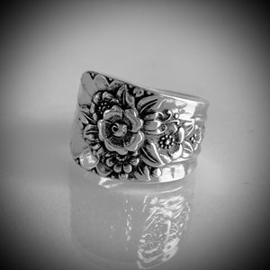 May include: A wide silver-toned ring featuring a prominent embossed floral pattern across the front surface. The design displays detailed petals and leaves with darkened recessed areas that highlight the intricate relief of the metalwork.