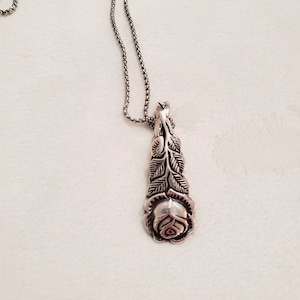 May include: A silver-toned necklace featuring a pendant shaped like a rose. The pendant has detailed leaf and petal designs. The chain is a delicate, linked style. The necklace is displayed on a soft, white surface.