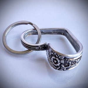 May include: A silver spoon keychain with a floral design. The spoon is bent into a square shape and has a silver ring attached to it.