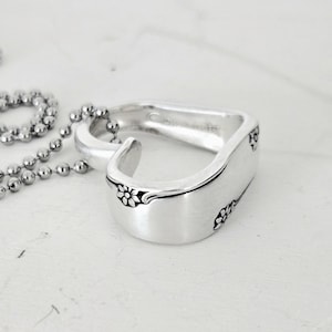 May include: A silver spoon ring with a floral design on the edges. The ring is connected to a silver ball chain necklace. The ring is on a white surface.
