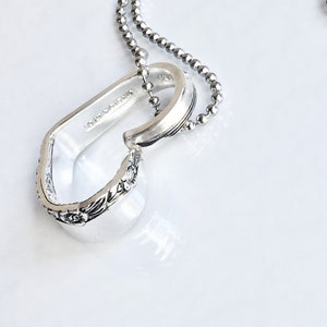 May include: A silver pendant necklace with a unique design. The pendant is crafted from a repurposed spoon, featuring floral detailing and a curved shape. The necklace has a ball chain. The background is white.
