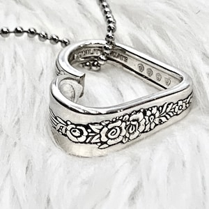 May include: A silver heart-shaped pendant necklace. The pendant is crafted from a spoon, featuring floral detailing and the word "PLATE". The necklace has a ball chain.