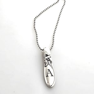 May include: Silver-toned pendant necklace featuring a spoon-shaped pendant with floral detailing and the letter "A". The pendant hangs from a ball chain necklace. The background is a plain white surface.