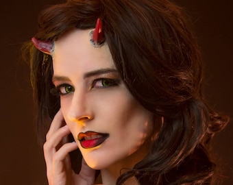May include: A woman with long dark hair and red horns on her forehead. She is wearing dark eye makeup with green eyeshadow and red lipstick. She is looking to the side and has her hand on her cheek.