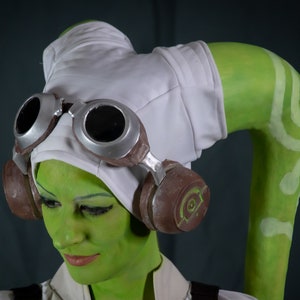 May include: A green-skinned person wearing a white hat with goggles and brown headphones. The person has a green and white striped tail.