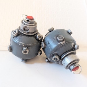 May include: Two gray, spherical decorative objects with silver accents and red tops. Each has a ring at the top and several protruding silver bolts. The objects are likely handmade and have a vintage or industrial aesthetic.