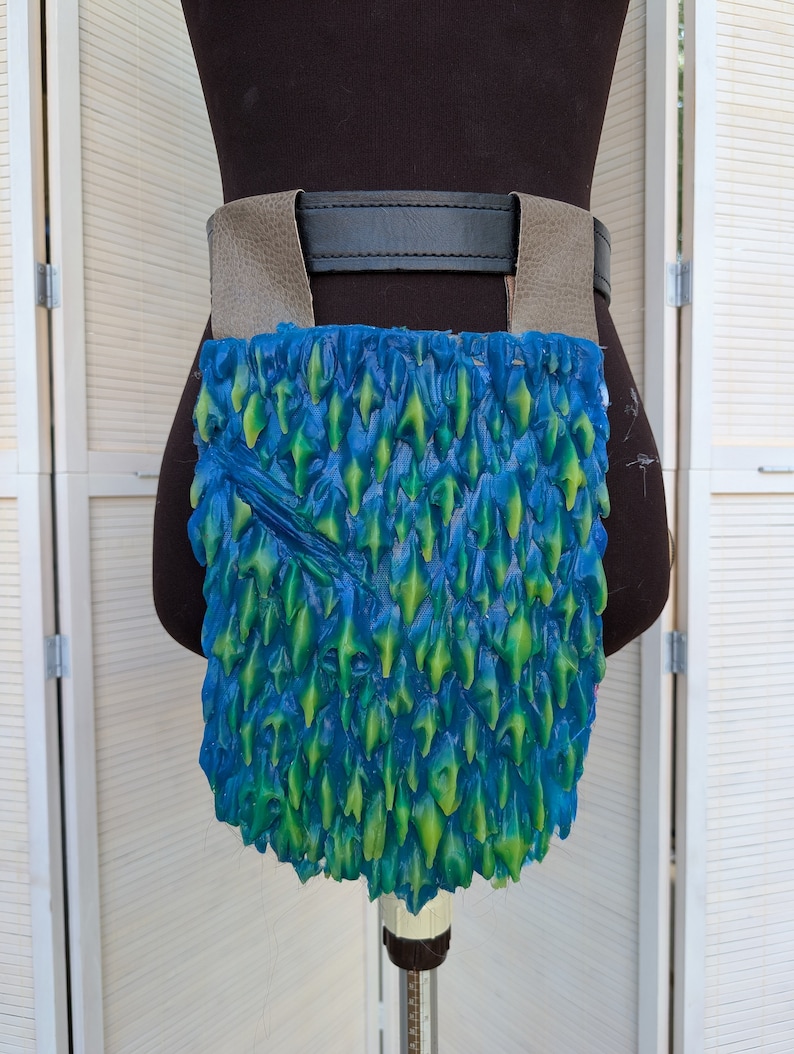 May include: A blue and green leather belt pouch with a scaly texture. The pouch is attached to a brown leather belt with a black buckle.