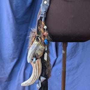 Op de afbeelding: Een zwarte leren riem met een bruine leren band en een metalen gesp. Een lange zwarte kwast hangt aan de riem met een verscheidenheid aan bedels, waaronder een nep dierentand, kralen en een kleine metalen bel.