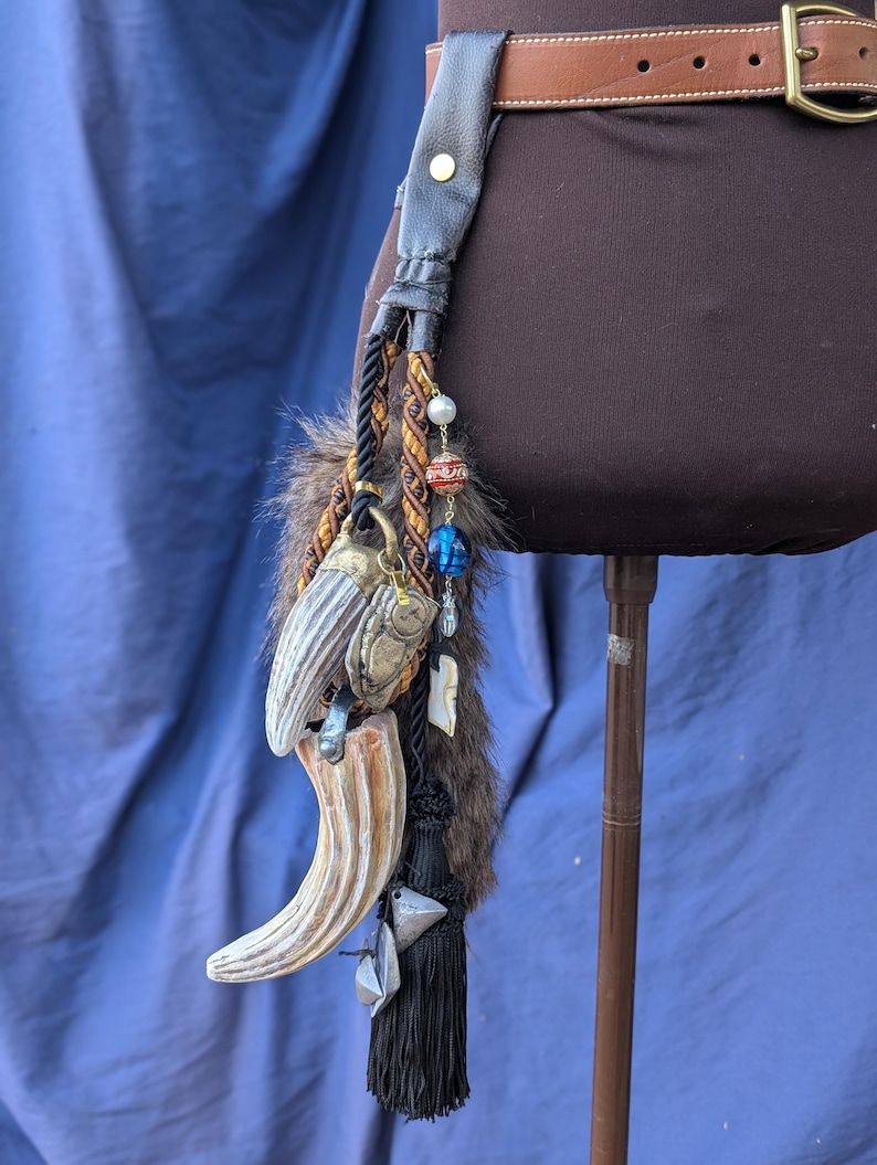Op de afbeelding: Een zwarte leren riem met een bruine leren band en een metalen gesp. Een lange zwarte kwast hangt aan de riem met een verscheidenheid aan bedels, waaronder een nep dierentand, kralen en een kleine metalen bel.