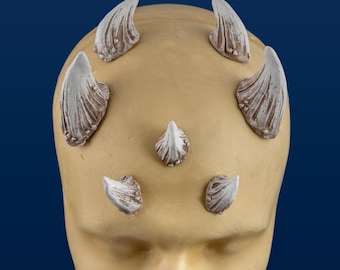 May include: Six white, sculpted horns are attached to a tan-colored mannequin head. The horns are various sizes and shapes, with some being more pointed than others.