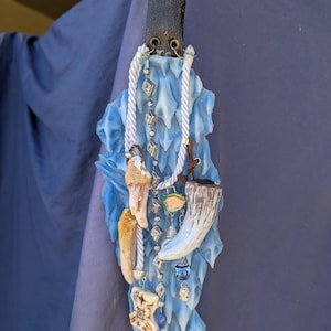 Op de afbeelding: Een blauwe en witte keramische wandhanger met verschillende bedels en kralen. De bedels omvatten een bot, een hoorn en een klein glazen flesje. De kralen zijn zilver, wit en blauw.