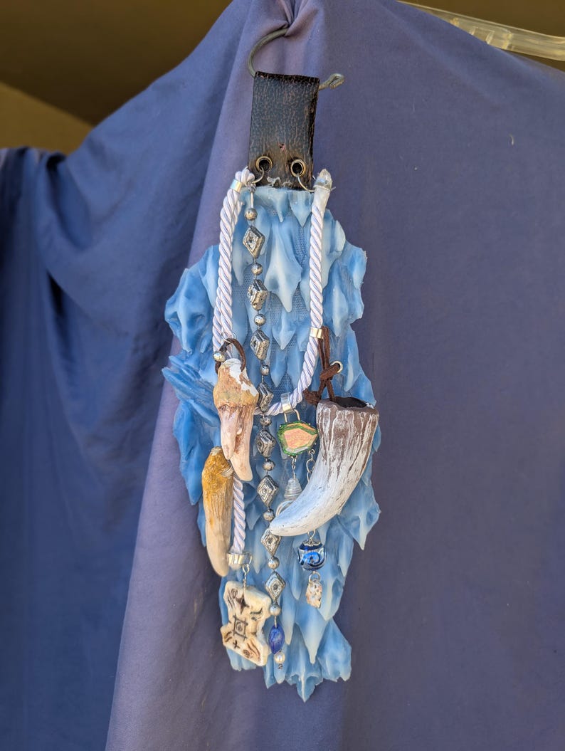 Op de afbeelding: Een blauwe en witte keramische wandhanger met verschillende bedels en kralen. De bedels omvatten een bot, een hoorn en een klein glazen flesje. De kralen zijn zilver, wit en blauw.