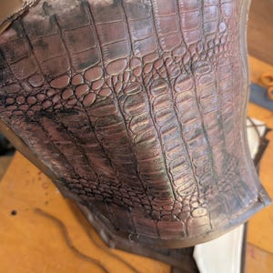 May include: Close-up of a brown leather piece with a crocodile-embossed pattern. The leather has a slightly distressed look, with some darker areas.