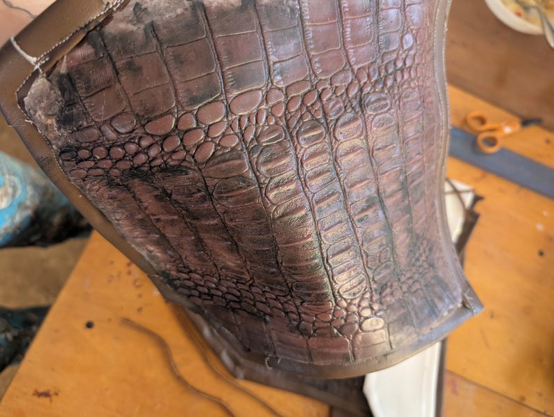 May include: Close-up of a brown leather piece with a crocodile-embossed pattern. The leather has a slightly distressed look, with some darker areas.