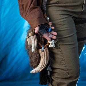 May include: A brown and white antler pendant with a fur accent, adorned with decorative beads and charms. The pendant is held against a blue backdrop, showcasing the intricate details and textures of the handcrafted item.