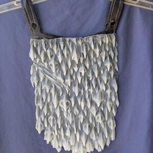 May include: A grey and white costume piece with a textured, spiky surface. The piece has two black leather straps with a button closure.
