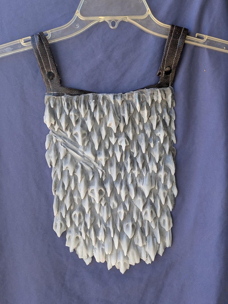 May include: A grey and white costume piece with a textured, spiky surface. The piece has two black leather straps with a button closure.