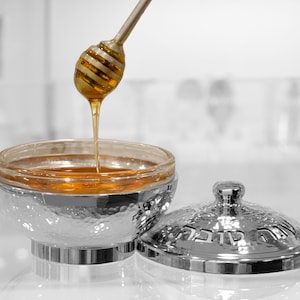 May include: A silver honey pot with a hammered finish and a matching lid. The lid has Hebrew writing on it. Honey is being poured from a honey dipper into the pot.